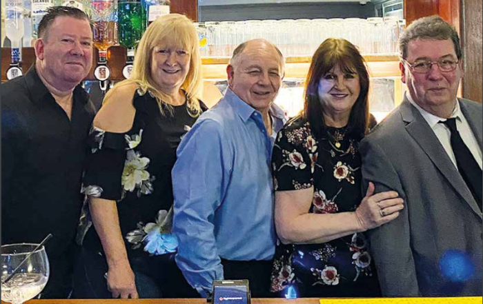 Five adults standing together and smiling at a bar, with glasses and bottles visible in the background.