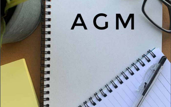 Spiral notebook with AGM on the cover, next to a pen, glasses, sticky notes, and a potted plant.