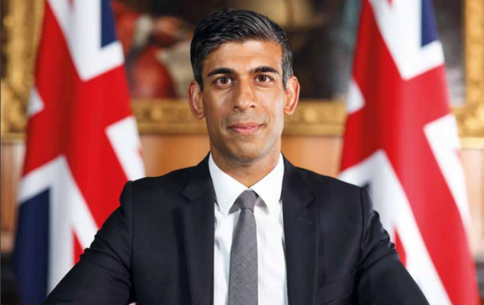 Rishi Sunak in a suit sits in front of two British flags with a neutral expression.