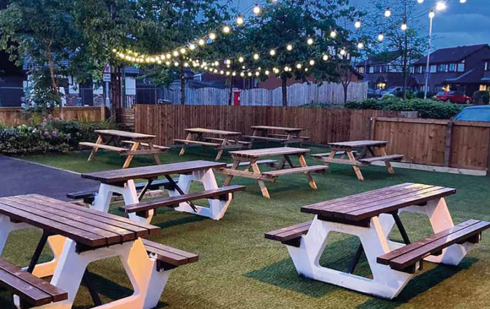 Outdoor seating with wooden picnic tables on grass, string lights overhead, and trees in the background.