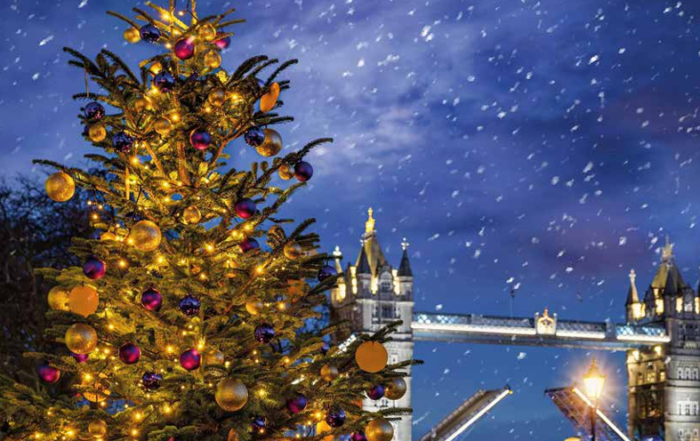 Christmas tree with lights and ornaments in front of Tower Bridge at night with falling snow.