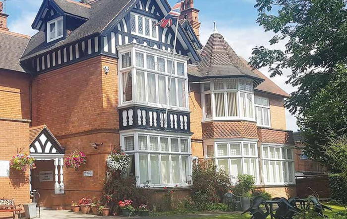 A large brick and timber Tudor-style house with gardens, a driveway, and a Union Jack flag on top.