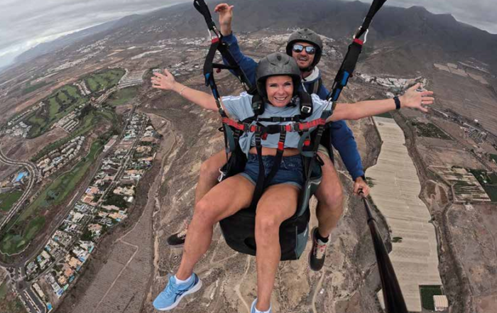 Two people paragliding above a landscape, with one smiling and arms outstretched while the other controls the glide.