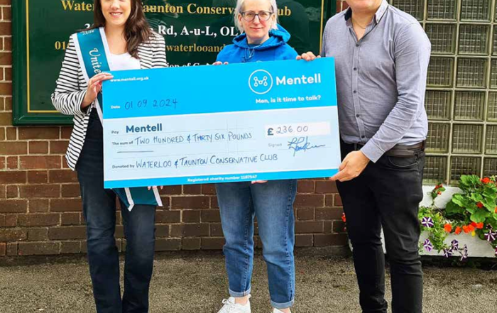 Three people holding a large blue check in front of a building with a sign.