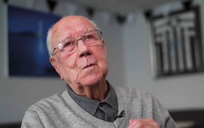 An elderly man wearing glasses and a gray sweater sits indoors, looking slightly upward.