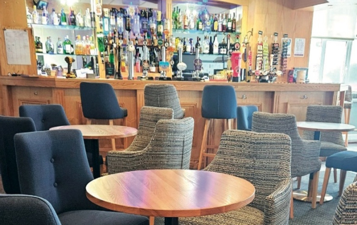 Modern lounge bar with empty round tables, cushioned chairs, and a stocked bar counter in the background.