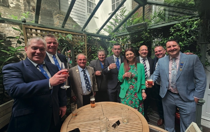 Nine people in formal attire stand around a table with drinks, posing for a group photo in a glass-roofed garden area.