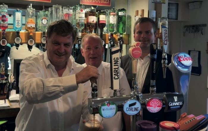 Three men in white shirts stand behind a bar with various beer taps and bottles of spirits in the background.