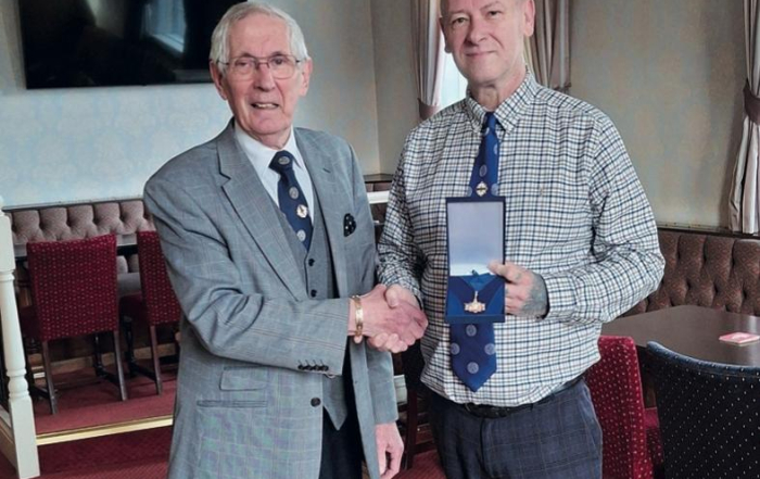 Two men shaking hands; one is holding an open box containing a medal in a formal indoor setting.