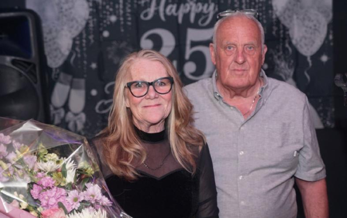 Two older adults standing together at a 25th anniversary celebration, one holding a bouquet of flowers.