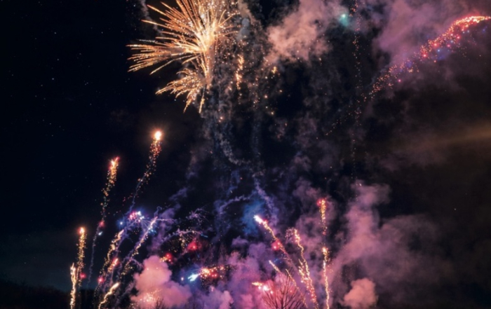 A crowd watches colorful fireworks explode in the night sky, with smoke and bright lights filling the scene.