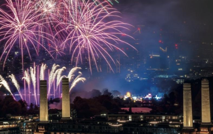 Fireworks light up the night sky above a cityscape with industrial buildings and distant illuminated structures.