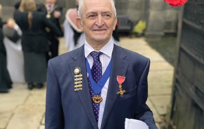 Man in a suit with medals stands outside, holding papers, with a red poppy in the air above him.